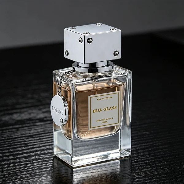 A lineup of three thick-bottomed rectangular clear glass perfume bottles containing peach, amber, and light green liquids. Each bottle is topped with a distinct cube-shaped cap accented with small rhinestone studs at the corners and a delicate metal hanging chain with a circular tag attached to the neck. The caps are finished in polished metallic silver, polished metallic gold, and solid black.