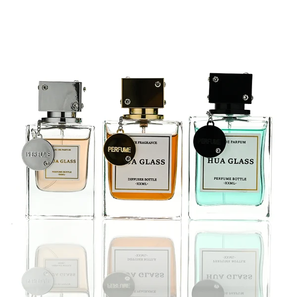 A lineup of three thick-bottomed rectangular clear glass perfume bottles containing peach, amber, and light green liquids. Each bottle is topped with a distinct cube-shaped cap accented with small rhinestone studs at the corners and a delicate metal hanging chain with a circular tag attached to the neck. The caps are finished in polished metallic silver, polished metallic gold, and solid black.