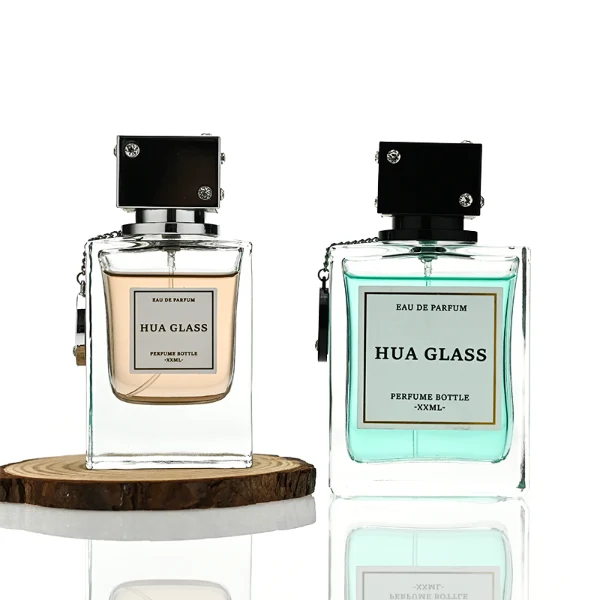 A lineup of three thick-bottomed rectangular clear glass perfume bottles containing peach, amber, and light green liquids. Each bottle is topped with a distinct cube-shaped cap accented with small rhinestone studs at the corners and a delicate metal hanging chain with a circular tag attached to the neck. The caps are finished in polished metallic silver, polished metallic gold, and solid black.