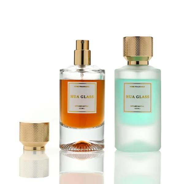 Two cylindrical glass perfume bottles: the left is finished in frosted white glass holding a green liquid, and the right is clear glass with a sunburst-grooved base holding an amber liquid. Both are paired with a heavy cylindrical metallic gold Zamac cap textured with a micro-diamond knurled pattern.