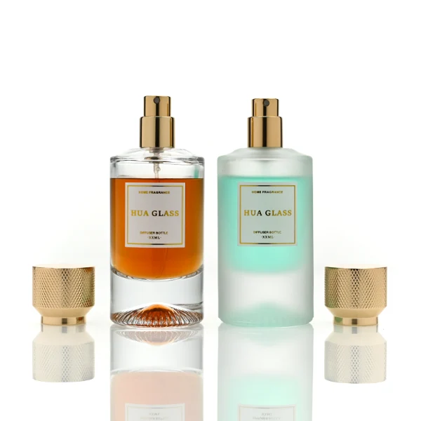 Two cylindrical glass perfume bottles: the left is finished in frosted white glass holding a green liquid, and the right is clear glass with a sunburst-grooved base holding an amber liquid. Both are paired with a heavy cylindrical metallic gold Zamac cap textured with a micro-diamond knurled pattern.