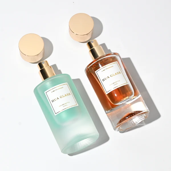 Two cylindrical glass perfume bottles: the left is finished in frosted white glass holding a green liquid, and the right is clear glass with a sunburst-grooved base holding an amber liquid. Both are paired with a heavy cylindrical metallic gold Zamac cap textured with a micro-diamond knurled pattern.