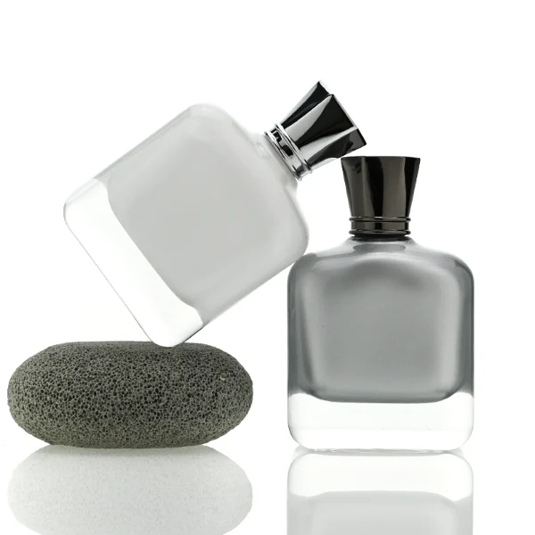 Two smooth rectangular glass perfume bottles with rounded corners and thick clear bases. One features an inner-sprayed metallic silver finish with a gunmetal black multifaceted geometric cap, while the other features an inner-sprayed opaque white finish topped with a chrome silver geometric cap.