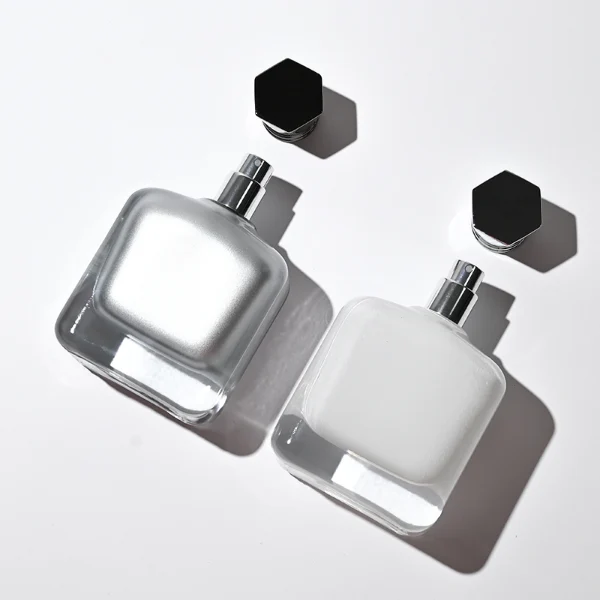 Two smooth rectangular glass perfume bottles with rounded corners and thick clear bases. One features an inner-sprayed metallic silver finish with a gunmetal black multifaceted geometric cap, while the other features an inner-sprayed opaque white finish topped with a chrome silver geometric cap.