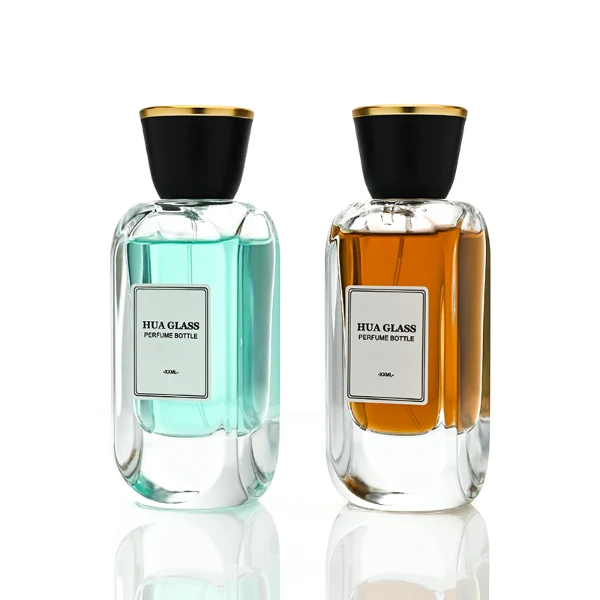 Two tall, thick-bottomed rectangular clear glass perfume bottles with rounded corners, each featuring a custom branded logo label on the front panel and filled with premium fragrance liquid. Both bottles are topped with a unique trumpet-shaped cap, which features a polished gloss black side body and a flat circular top completely covered in a polished metallic gold layer.