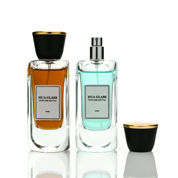 Two tall, thick-bottomed rectangular clear glass perfume bottles with rounded corners, each featuring a custom branded logo label on the front panel and filled with premium fragrance liquid. Both bottles are topped with a unique trumpet-shaped cap, which features a polished gloss black side body and a flat circular top completely covered in a polished metallic gold layer.