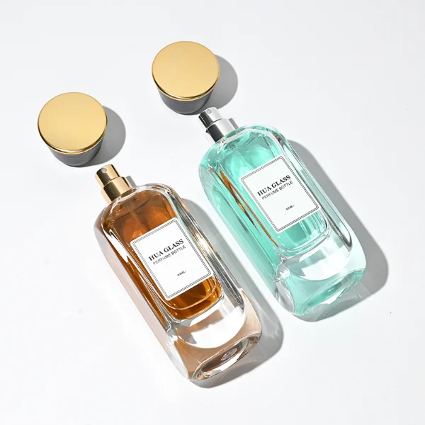 Two tall, thick-bottomed rectangular clear glass perfume bottles with rounded corners, each featuring a custom branded logo label on the front panel and filled with premium fragrance liquid. Both bottles are topped with a unique trumpet-shaped cap, which features a polished gloss black side body and a flat circular top completely covered in a polished metallic gold layer.