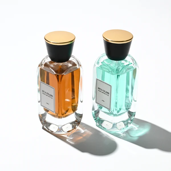Two tall, thick-bottomed rectangular clear glass perfume bottles with rounded corners, each featuring a custom branded logo label on the front panel and filled with premium fragrance liquid. Both bottles are topped with a unique trumpet-shaped cap, which features a polished gloss black side body and a flat circular top completely covered in a polished metallic gold layer.