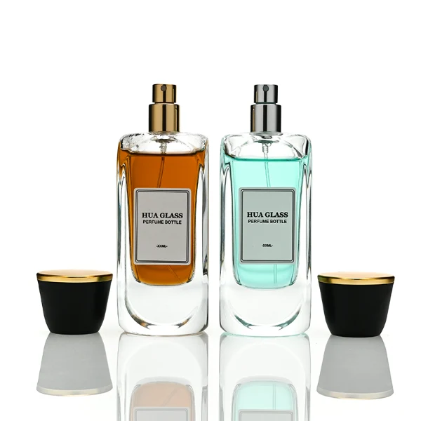Two tall, thick-bottomed rectangular clear glass perfume bottles with rounded corners, each featuring a custom branded logo label on the front panel and filled with premium fragrance liquid. Both bottles are topped with a unique trumpet-shaped cap, which features a polished gloss black side body and a flat circular top completely covered in a polished metallic gold layer.