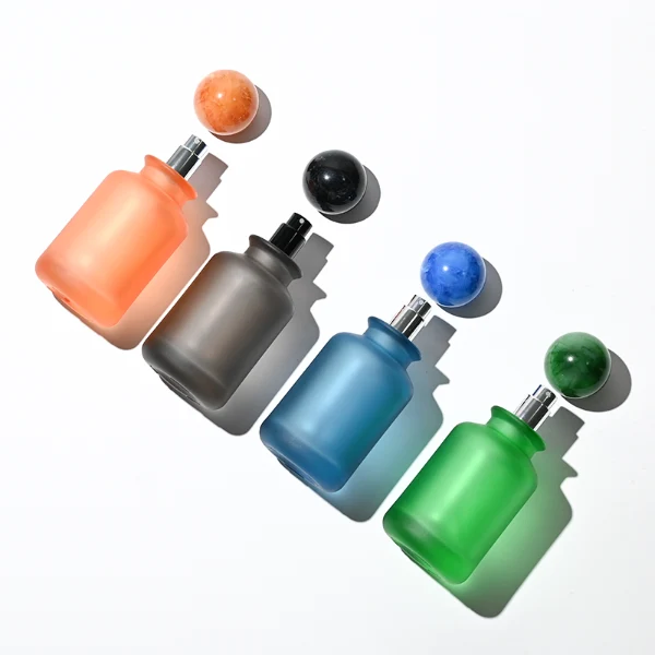 A lineup of four cylindrical frosted glass perfume bottles in matte shades of blue, orange, dark grey, and green. Each bottle is paired with a matching large spherical ball cap featuring a glossy, marbled stone texture in deep blue, amber brown, black, and forest green.