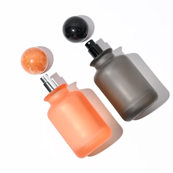 A lineup of four cylindrical frosted glass perfume bottles in matte shades of blue, orange, dark grey, and green. Each bottle is paired with a matching large spherical ball cap featuring a glossy, marbled stone texture in deep blue, amber brown, black, and forest green.