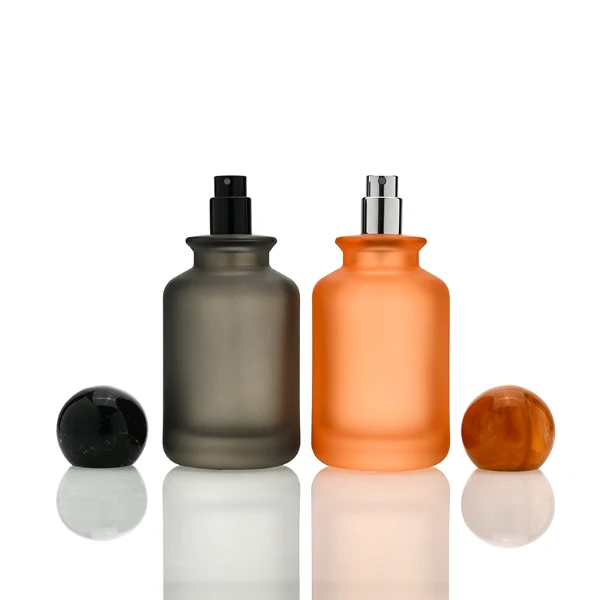 A lineup of four cylindrical frosted glass perfume bottles in matte shades of blue, orange, dark grey, and green. Each bottle is paired with a matching large spherical ball cap featuring a glossy, marbled stone texture in deep blue, amber brown, black, and forest green.