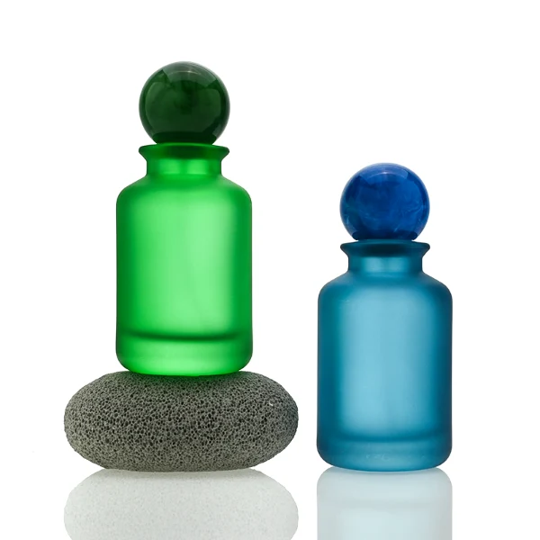 A lineup of four cylindrical frosted glass perfume bottles in matte shades of blue, orange, dark grey, and green. Each bottle is paired with a matching large spherical ball cap featuring a glossy, marbled stone texture in deep blue, amber brown, black, and forest green.