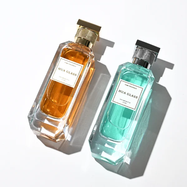 Two tall, thick-bottomed rectangular clear glass perfume bottles with beveled corners, containing amber and light green liquids. Both bottles feature an inset front panel for labeling and are paired with unique cylindrical caps featuring a textured diamond-knurled collar and a smooth, flat square top, shown in polished gold and metallic silver finishes.