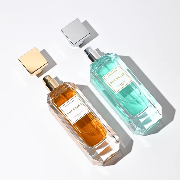 Two tall, thick-bottomed rectangular clear glass perfume bottles with beveled corners, containing amber and light green liquids. Both bottles feature an inset front panel for labeling and are paired with unique cylindrical caps featuring a textured diamond-knurled collar and a smooth, flat square top, shown in polished gold and metallic silver finishes.