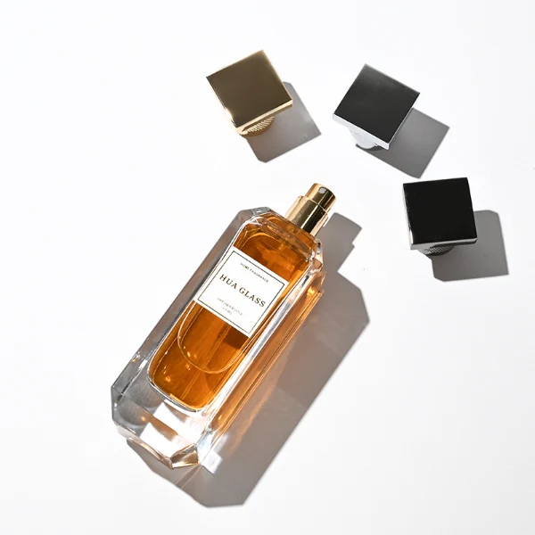 Two tall, thick-bottomed rectangular clear glass perfume bottles with beveled corners, containing amber and light green liquids. Both bottles feature an inset front panel for labeling and are paired with unique cylindrical caps featuring a textured diamond-knurled collar and a smooth, flat square top, shown in polished gold and metallic silver finishes.