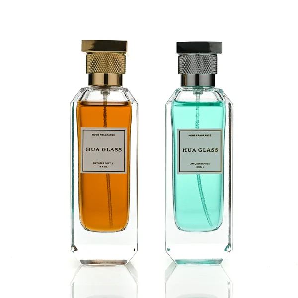 Two tall, thick-bottomed rectangular clear glass perfume bottles with beveled corners, containing amber and light green liquids. Both bottles feature an inset front panel for labeling and are paired with unique cylindrical caps featuring a textured diamond-knurled collar and a smooth, flat square top, shown in polished gold and metallic silver finishes.