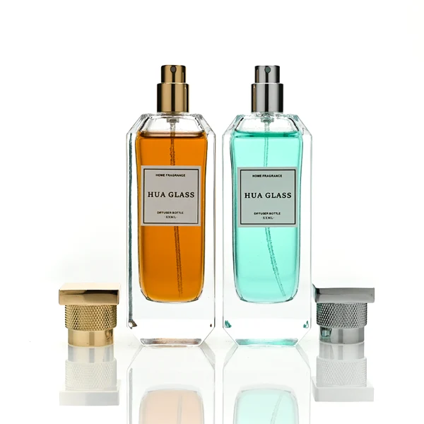 Two tall, thick-bottomed rectangular clear glass perfume bottles with beveled corners, containing amber and light green liquids. Both bottles feature an inset front panel for labeling and are paired with unique cylindrical caps featuring a textured diamond-knurled collar and a smooth, flat square top, shown in polished gold and metallic silver finishes.