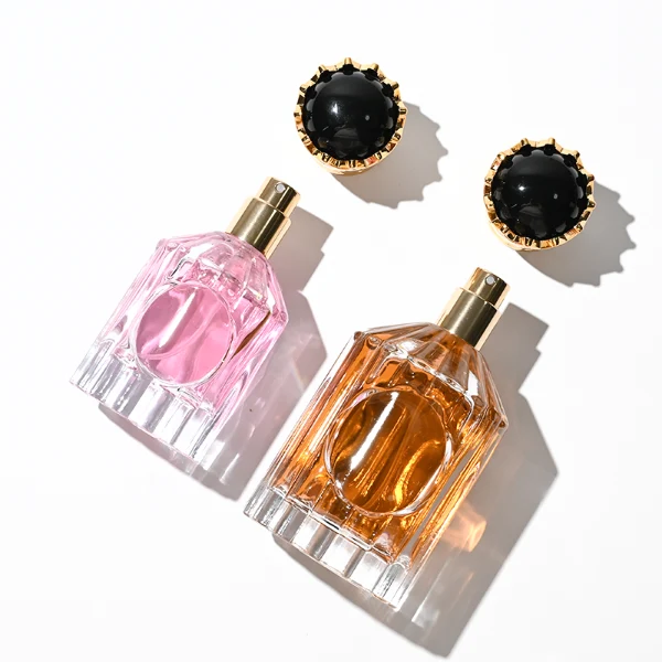 Two clear glass perfume bottles with vertical ridges, sloped shoulders, and a smooth circular inset on the front panel, holding amber and vibrant pink liquids. Both bottles are crowned with ornate, high-shine golden crown-motif caps topped with a glossy black domed center.