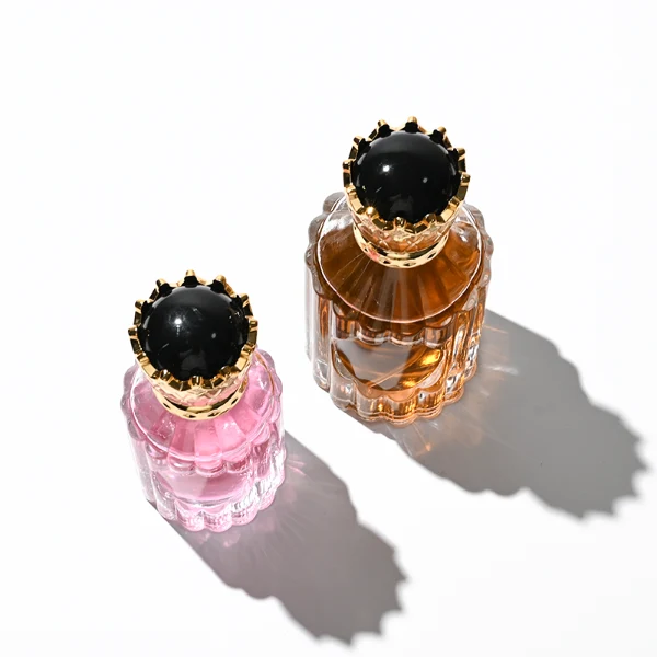 Two clear glass perfume bottles with vertical ridges, sloped shoulders, and a smooth circular inset on the front panel, holding amber and vibrant pink liquids. Both bottles are crowned with ornate, high-shine golden crown-motif caps topped with a glossy black domed center.