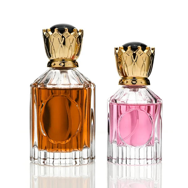Two clear glass perfume bottles with vertical ridges, sloped shoulders, and a smooth circular inset on the front panel, holding amber and vibrant pink liquids. Both bottles are crowned with ornate, high-shine golden crown-motif caps topped with a glossy black domed center.