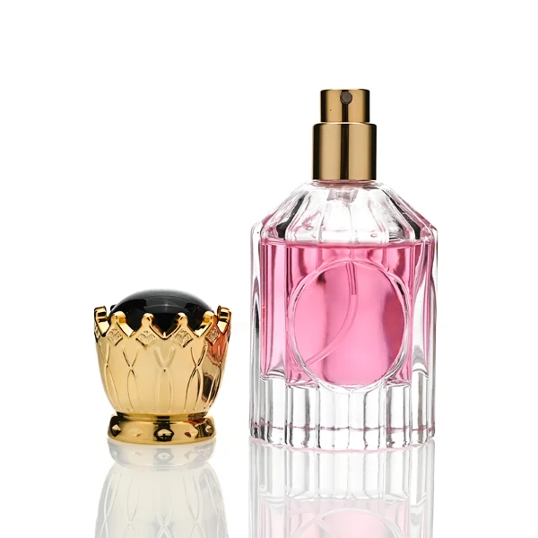 Two clear glass perfume bottles with vertical ridges, sloped shoulders, and a smooth circular inset on the front panel, holding amber and vibrant pink liquids. Both bottles are crowned with ornate, high-shine golden crown-motif caps topped with a glossy black domed center.