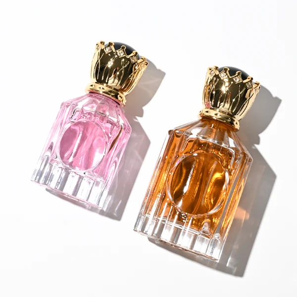 Two clear glass perfume bottles with vertical ridges, sloped shoulders, and a smooth circular inset on the front panel, holding amber and vibrant pink liquids. Both bottles are crowned with ornate, high-shine golden crown-motif caps topped with a glossy black domed center.