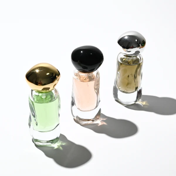 Three clear glass perfume bottles featuring smoothly curved organic shapes and heavy transparent bases, filled with olive green, pale pink, and light green liquids. The bottles are topped with smooth, pebble-like bulbous round caps, displayed in glossy black, polished metallic gold, and high-shine metallic silver finishes.