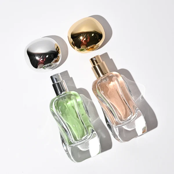 Three clear glass perfume bottles featuring smoothly curved organic shapes and heavy transparent bases, filled with olive green, pale pink, and light green liquids. The bottles are topped with smooth, pebble-like bulbous round caps, displayed in glossy black, polished metallic gold, and high-shine metallic silver finishes.