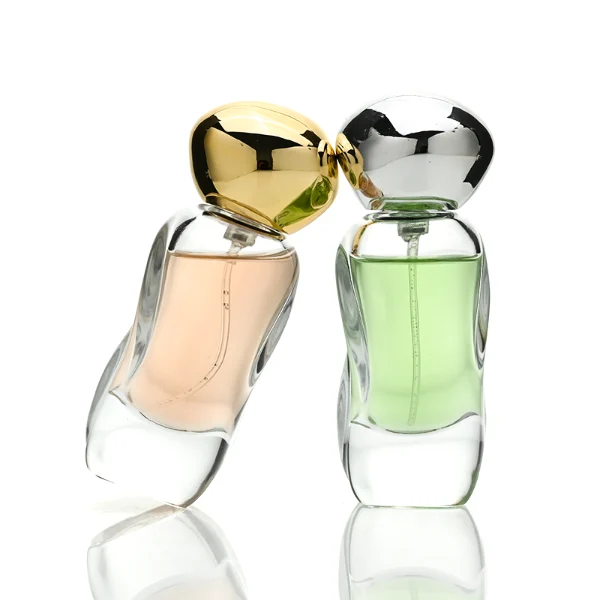 Three clear glass perfume bottles featuring smoothly curved organic shapes and heavy transparent bases, filled with olive green, pale pink, and light green liquids. The bottles are topped with smooth, pebble-like bulbous round caps, displayed in glossy black, polished metallic gold, and high-shine metallic silver finishes.