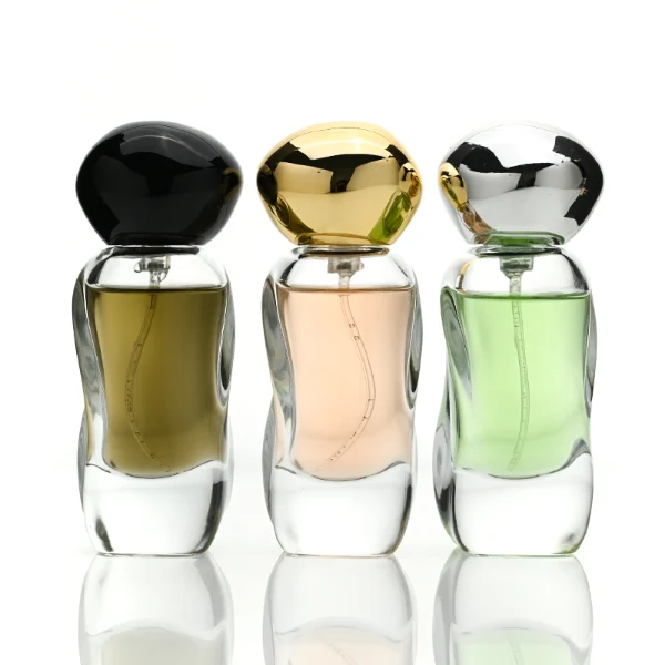 Three clear glass perfume bottles featuring smoothly curved organic shapes and heavy transparent bases, filled with olive green, pale pink, and light green liquids. The bottles are topped with smooth, pebble-like bulbous round caps, displayed in glossy black, polished metallic gold, and high-shine metallic silver finishes.