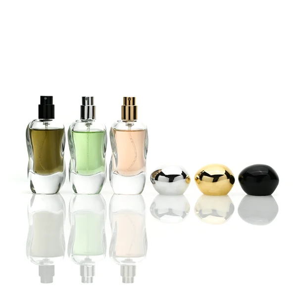Three clear glass perfume bottles featuring smoothly curved organic shapes and heavy transparent bases, filled with olive green, pale pink, and light green liquids. The bottles are topped with smooth, pebble-like bulbous round caps, displayed in glossy black, polished metallic gold, and high-shine metallic silver finishes.