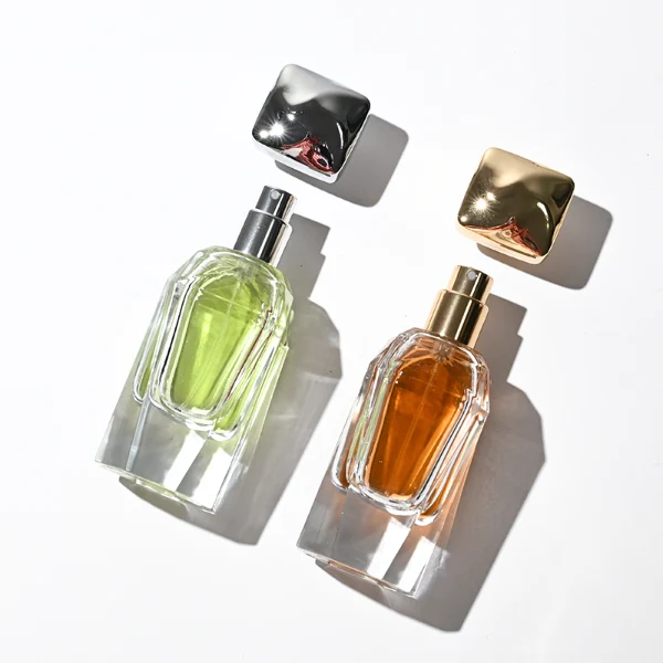 Two clear glass perfume bottles featuring prominent multi-faceted angular geometric sides and thick clear bases, holding light green and amber liquids. The bottles are topped with a heavy, polished Zamac cube cap with stepped detailing at the collar, shown in high-shine chrome silver and brilliant polished gold finishes.