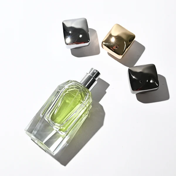 Two clear glass perfume bottles featuring prominent multi-faceted angular geometric sides and thick clear bases, holding light green and amber liquids. The bottles are topped with a heavy, polished Zamac cube cap with stepped detailing at the collar, shown in high-shine chrome silver and brilliant polished gold finishes.