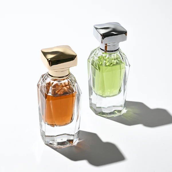 Two clear glass perfume bottles featuring prominent multi-faceted angular geometric sides and thick clear bases, holding light green and amber liquids. The bottles are topped with a heavy, polished Zamac cube cap with stepped detailing at the collar, shown in high-shine chrome silver and brilliant polished gold finishes.