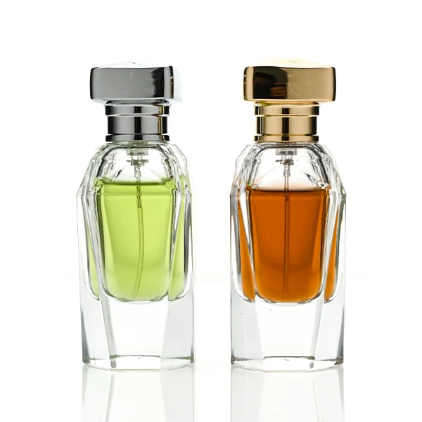 Two clear glass perfume bottles featuring prominent multi-faceted angular geometric sides and thick clear bases, holding light green and amber liquids. The bottles are topped with a heavy, polished Zamac cube cap with stepped detailing at the collar, shown in high-shine chrome silver and brilliant polished gold finishes.