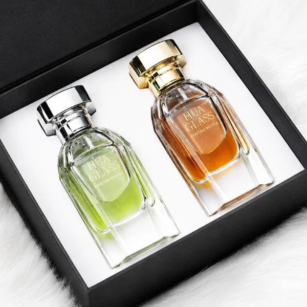 Two clear glass perfume bottles featuring prominent multi-faceted angular geometric sides and thick clear bases, holding light green and amber liquids. The bottles are topped with a heavy, polished Zamac cube cap with stepped detailing at the collar, shown in high-shine chrome silver and brilliant polished gold finishes.
