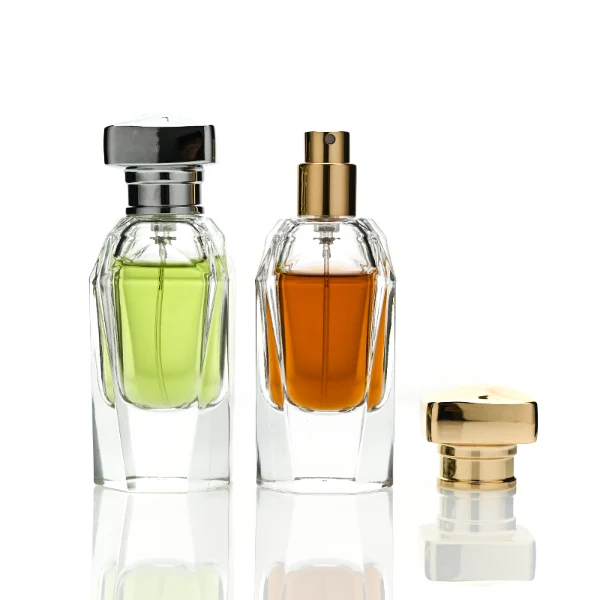 Two clear glass perfume bottles featuring prominent multi-faceted angular geometric sides and thick clear bases, holding light green and amber liquids. The bottles are topped with a heavy, polished Zamac cube cap with stepped detailing at the collar, shown in high-shine chrome silver and brilliant polished gold finishes.