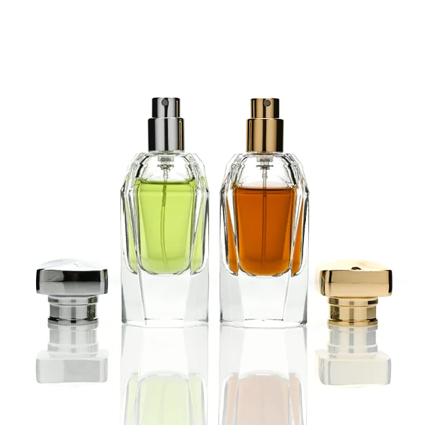 Two clear glass perfume bottles featuring prominent multi-faceted angular geometric sides and thick clear bases, holding light green and amber liquids. The bottles are topped with a heavy, polished Zamac cube cap with stepped detailing at the collar, shown in high-shine chrome silver and brilliant polished gold finishes.
