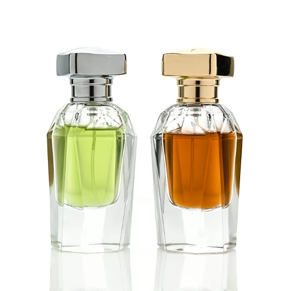 Two clear glass perfume bottles featuring prominent multi-faceted angular geometric sides and thick clear bases, holding light green and amber liquids. The bottles are topped with a heavy, polished Zamac cube cap with stepped detailing at the collar, shown in high-shine chrome silver and brilliant polished gold finishes.