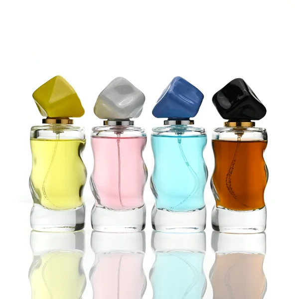 A lineup of four clear glass perfume bottles featuring unique, wave-curved organic sides and heavy transparent bases. The bottles are filled with yellow, pink, light blue, and amber liquids, each topped with an asymmetrical, abstract artistic cap resembling a tumbled stone or distorted cube in glossy mustard yellow, light grey, periwinkle blue, and solid black finishes.