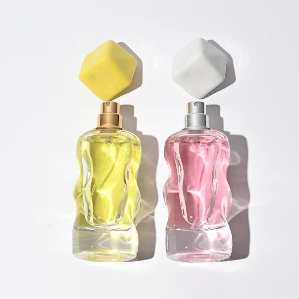 A lineup of four clear glass perfume bottles featuring unique, wave-curved organic sides and heavy transparent bases. The bottles are filled with yellow, pink, light blue, and amber liquids, each topped with an asymmetrical, abstract artistic cap resembling a tumbled stone or distorted cube in glossy mustard yellow, light grey, periwinkle blue, and solid black finishes.