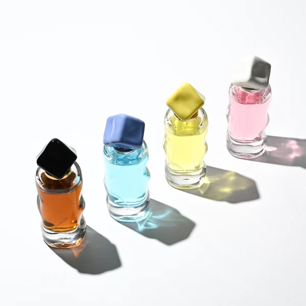 A lineup of four clear glass perfume bottles featuring unique, wave-curved organic sides and heavy transparent bases. The bottles are filled with yellow, pink, light blue, and amber liquids, each topped with an asymmetrical, abstract artistic cap resembling a tumbled stone or distorted cube in glossy mustard yellow, light grey, periwinkle blue, and solid black finishes.