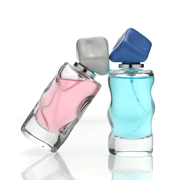 A lineup of four clear glass perfume bottles featuring unique, wave-curved organic sides and heavy transparent bases. The bottles are filled with yellow, pink, light blue, and amber liquids, each topped with an asymmetrical, abstract artistic cap resembling a tumbled stone or distorted cube in glossy mustard yellow, light grey, periwinkle blue, and solid black finishes.