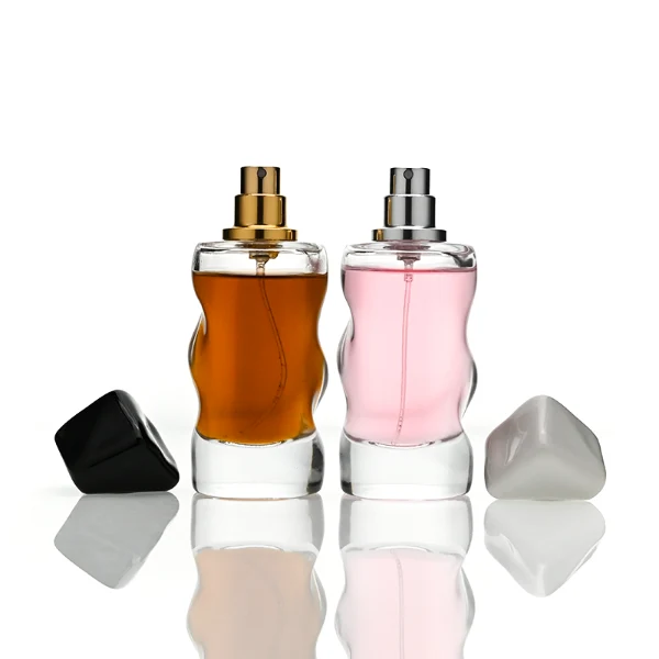 A lineup of four clear glass perfume bottles featuring unique, wave-curved organic sides and heavy transparent bases. The bottles are filled with yellow, pink, light blue, and amber liquids, each topped with an asymmetrical, abstract artistic cap resembling a tumbled stone or distorted cube in glossy mustard yellow, light grey, periwinkle blue, and solid black finishes.