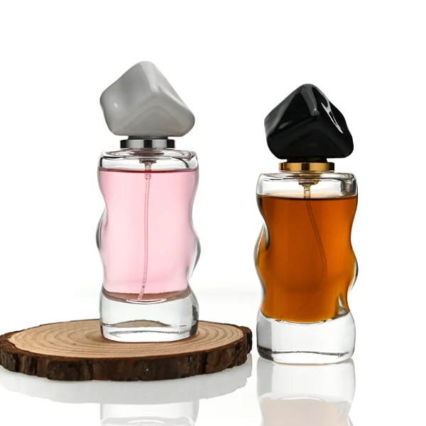 A lineup of four clear glass perfume bottles featuring unique, wave-curved organic sides and heavy transparent bases. The bottles are filled with yellow, pink, light blue, and amber liquids, each topped with an asymmetrical, abstract artistic cap resembling a tumbled stone or distorted cube in glossy mustard yellow, light grey, periwinkle blue, and solid black finishes.