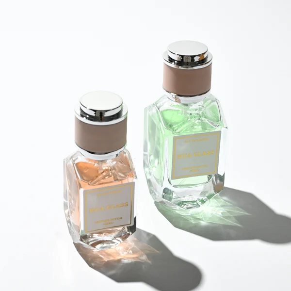 Two multi-faceted, gemstone-shaped clear glass perfume bottles with thick faceted bases, holding peach and light green liquids. Each bottle features a square white label and is topped with a unique cylindrical cap showcasing a matte beige center band paired with polished silver metallic trim.
