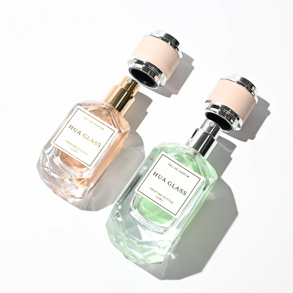 Two multi-faceted, gemstone-shaped clear glass perfume bottles with thick faceted bases, holding peach and light green liquids. Each bottle features a square white label and is topped with a unique cylindrical cap showcasing a matte beige center band paired with polished silver metallic trim.