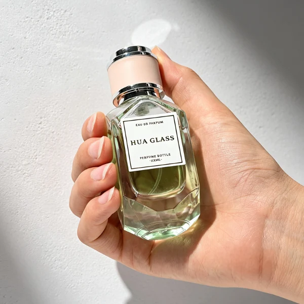 Two multi-faceted, gemstone-shaped clear glass perfume bottles with thick faceted bases, holding peach and light green liquids. Each bottle features a square white label and is topped with a unique cylindrical cap showcasing a matte beige center band paired with polished silver metallic trim.