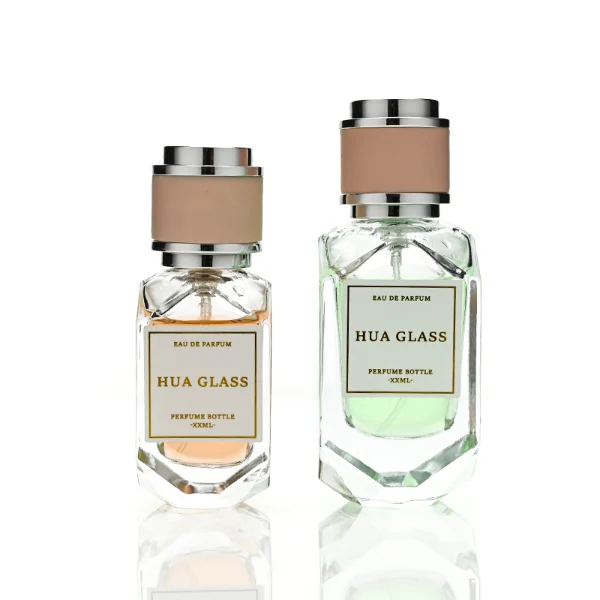 Two multi-faceted, gemstone-shaped clear glass perfume bottles with thick faceted bases, holding peach and light green liquids. Each bottle features a square white label and is topped with a unique cylindrical cap showcasing a matte beige center band paired with polished silver metallic trim.